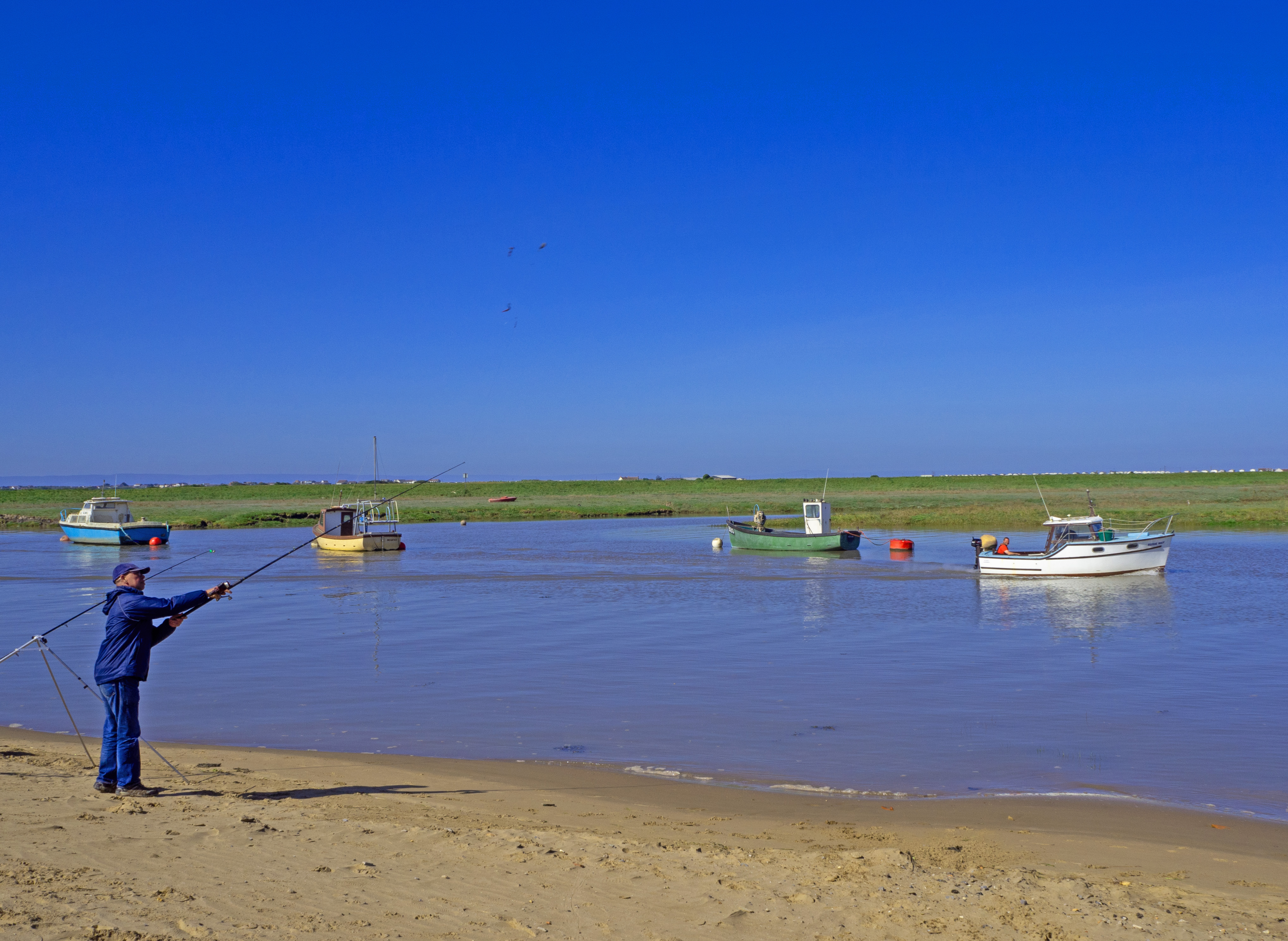 Fisherman with two fishing rods at high tide in Somreset with four boats on the river