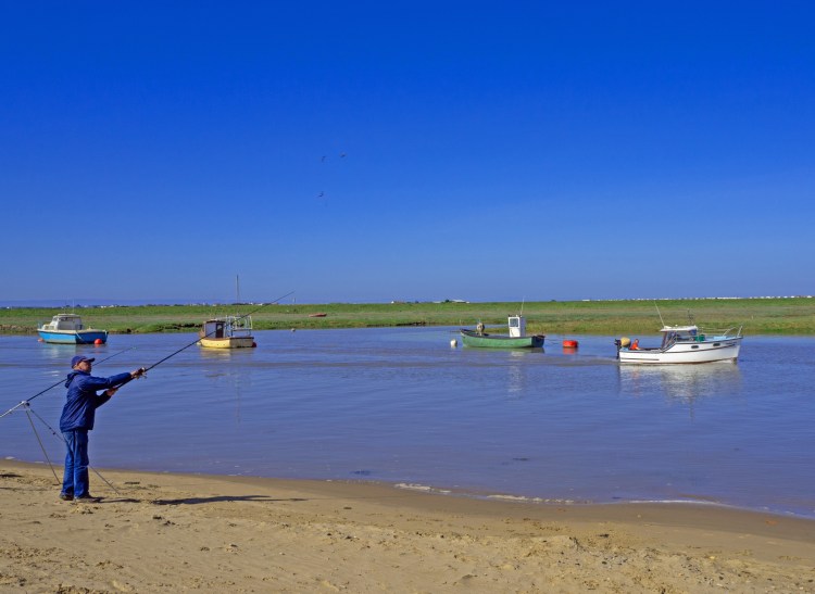 Fisherman with two fishing rods at high tide in Somreset with four boats on the river
