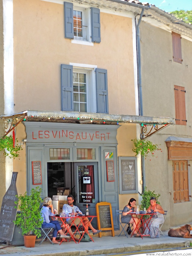people sat at tables drinking wine on a wine bar terrace in Provence with dog and old French buildings with shutters