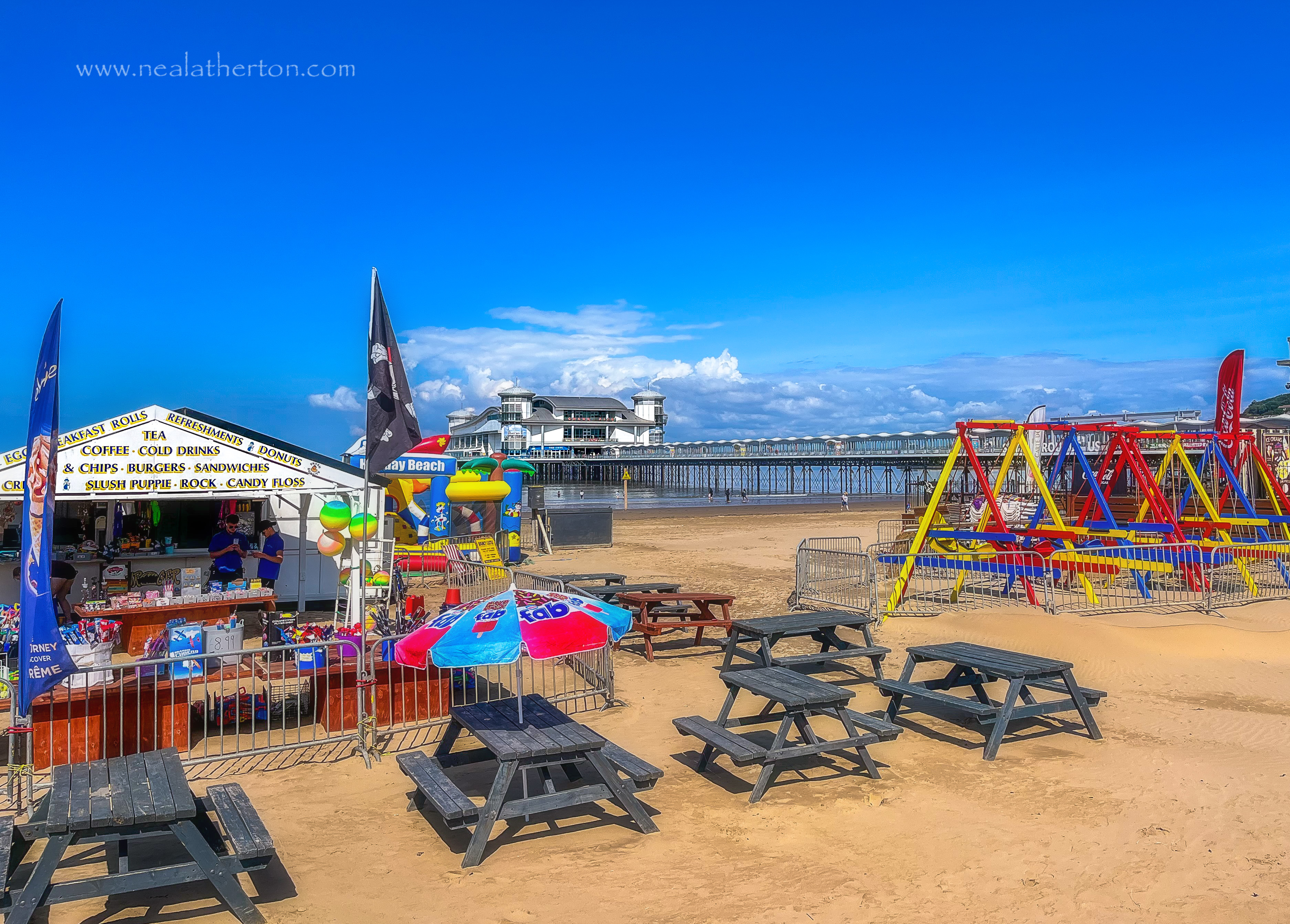 Seats and tables in front of refreshment stand with the GRand pier and swings to the side under blue summer sky