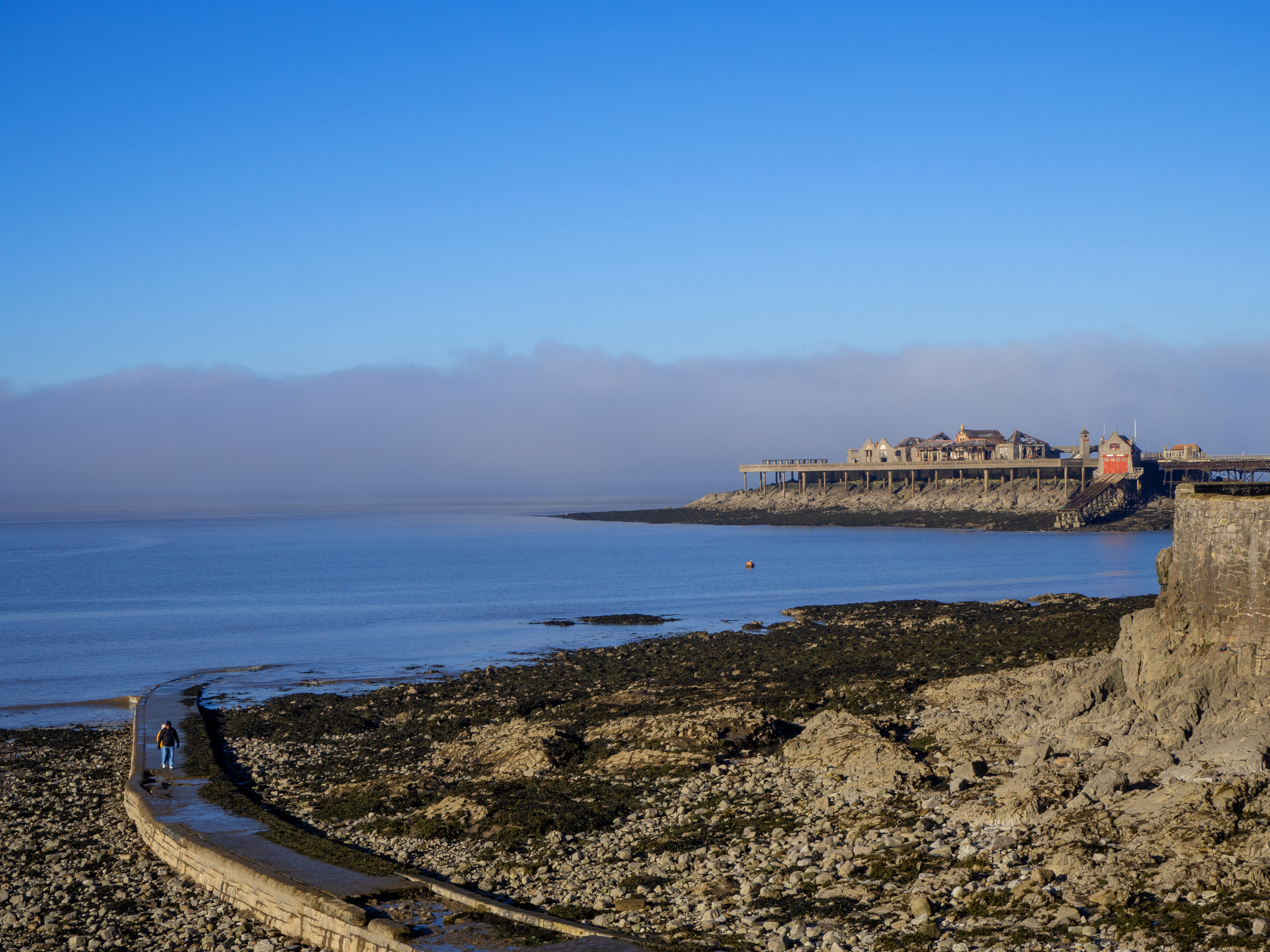 man walking on cove walkway low tide with blue sky and mist over pier and sea