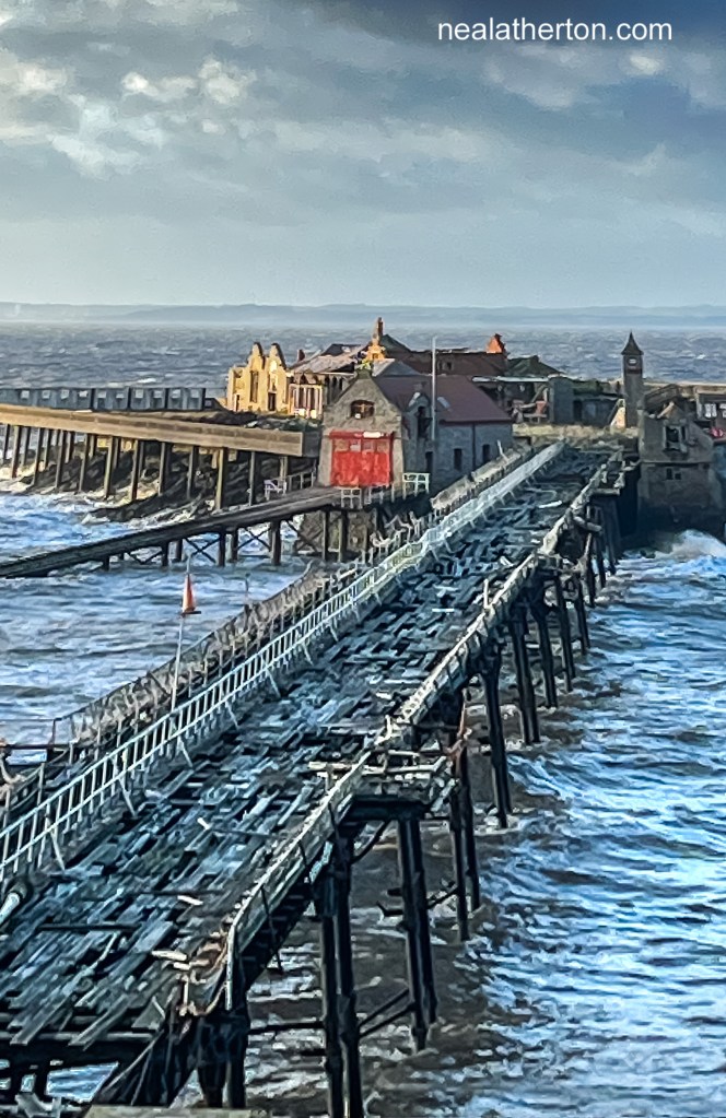 Old abandoned pier with much damage to walkway with RNLI station in front of ruined biuldings