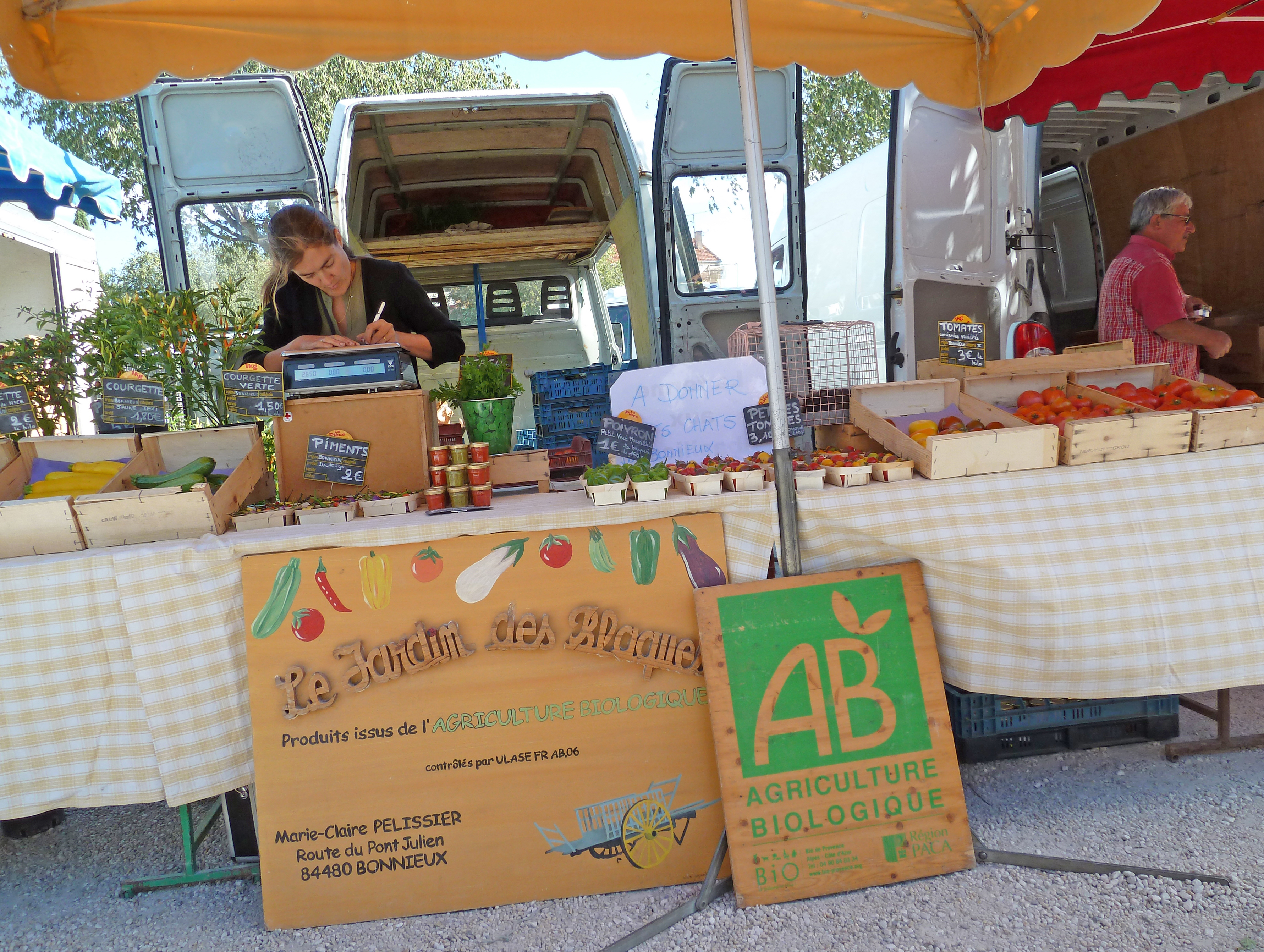 lady writing on fruit market stall with signs in front and van with man walking