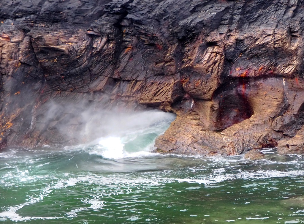 water rushes from hole in cliff into sea with high cliff and choppy water