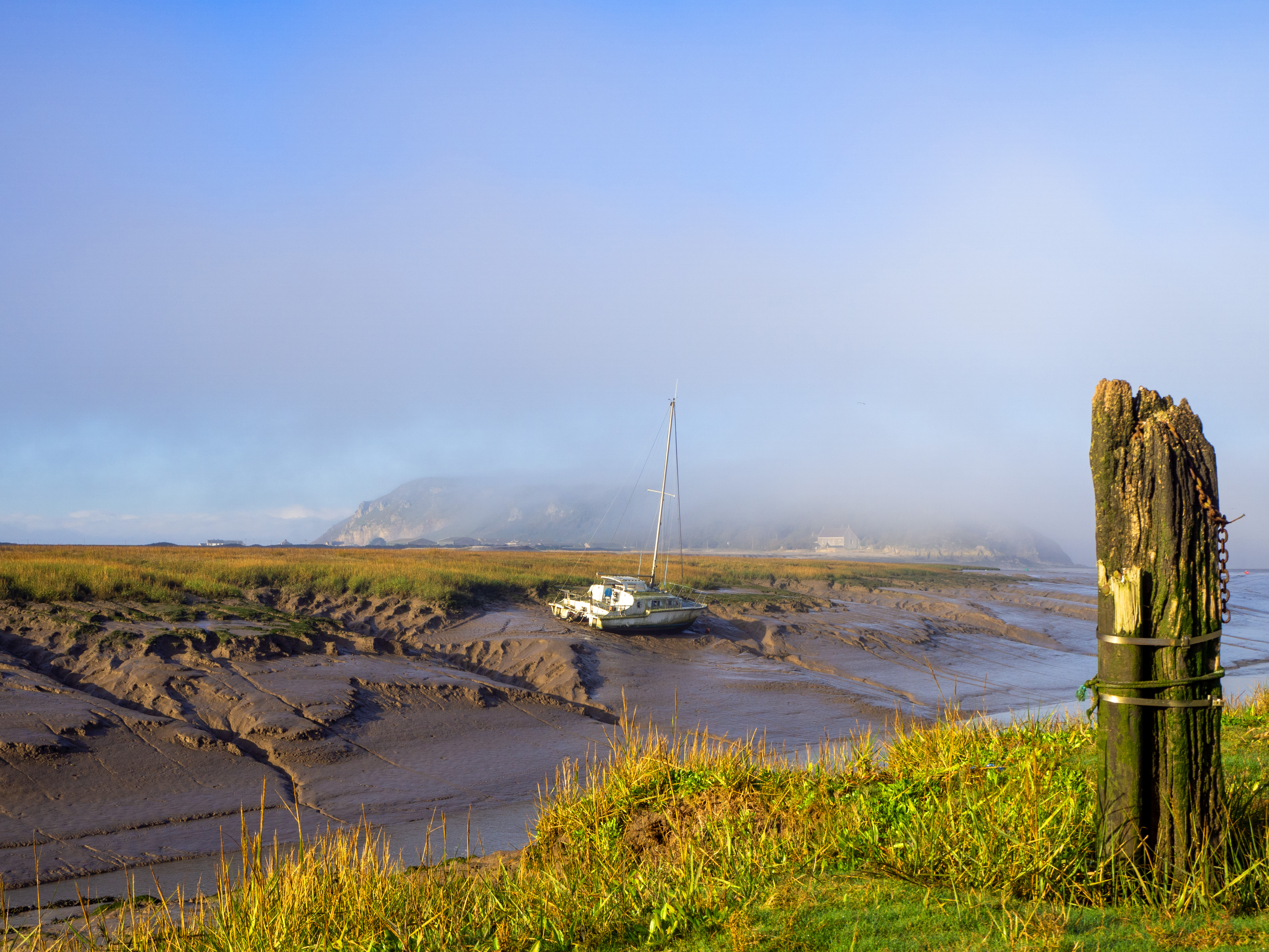 mooring post by river estuary with yacht on mud before island and mist in sky
