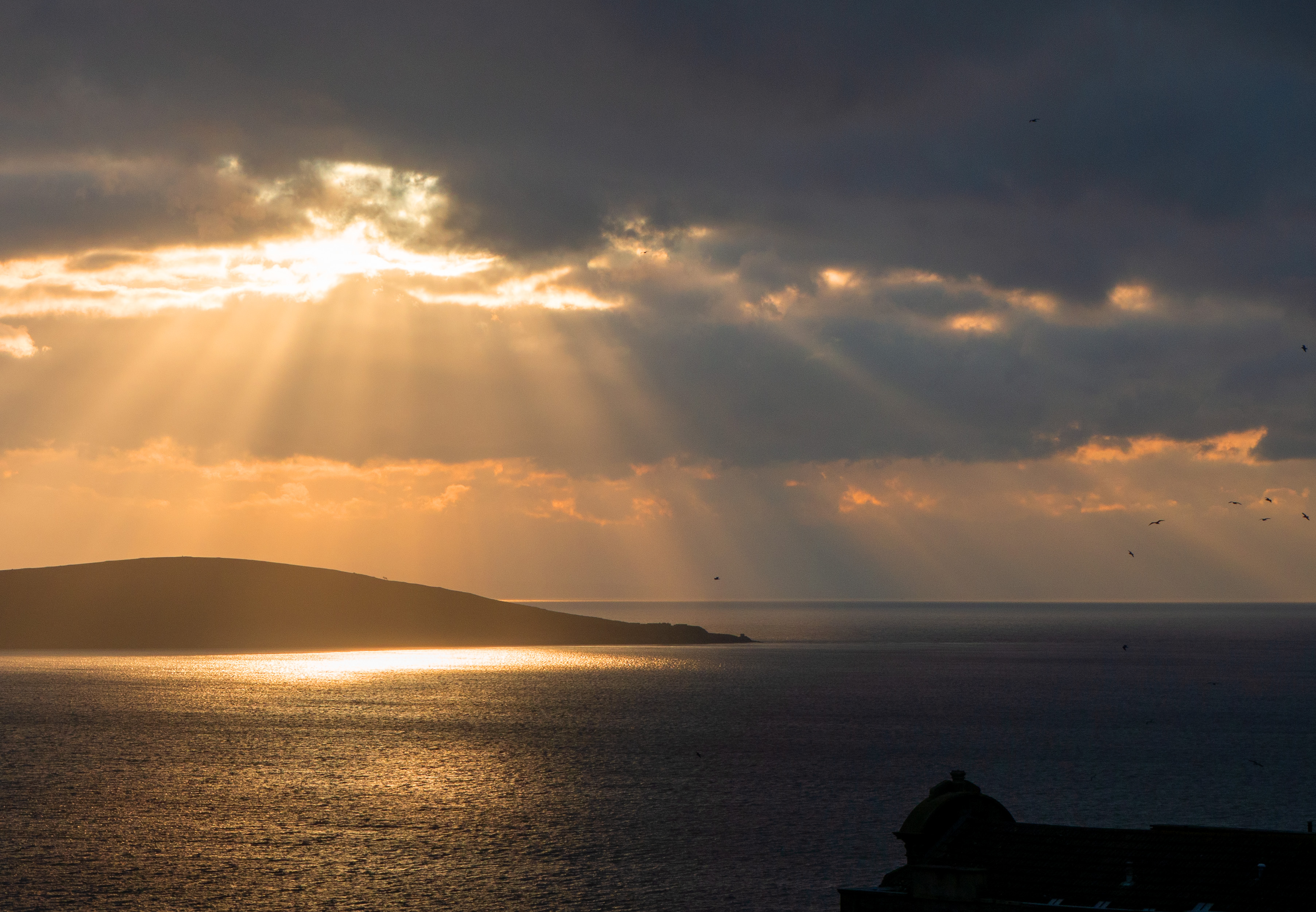 sun bursts through clouds onto sea with island and land in distance
