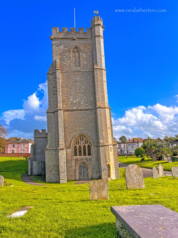 gravestones on grass in front of church tower with houses and blue sky