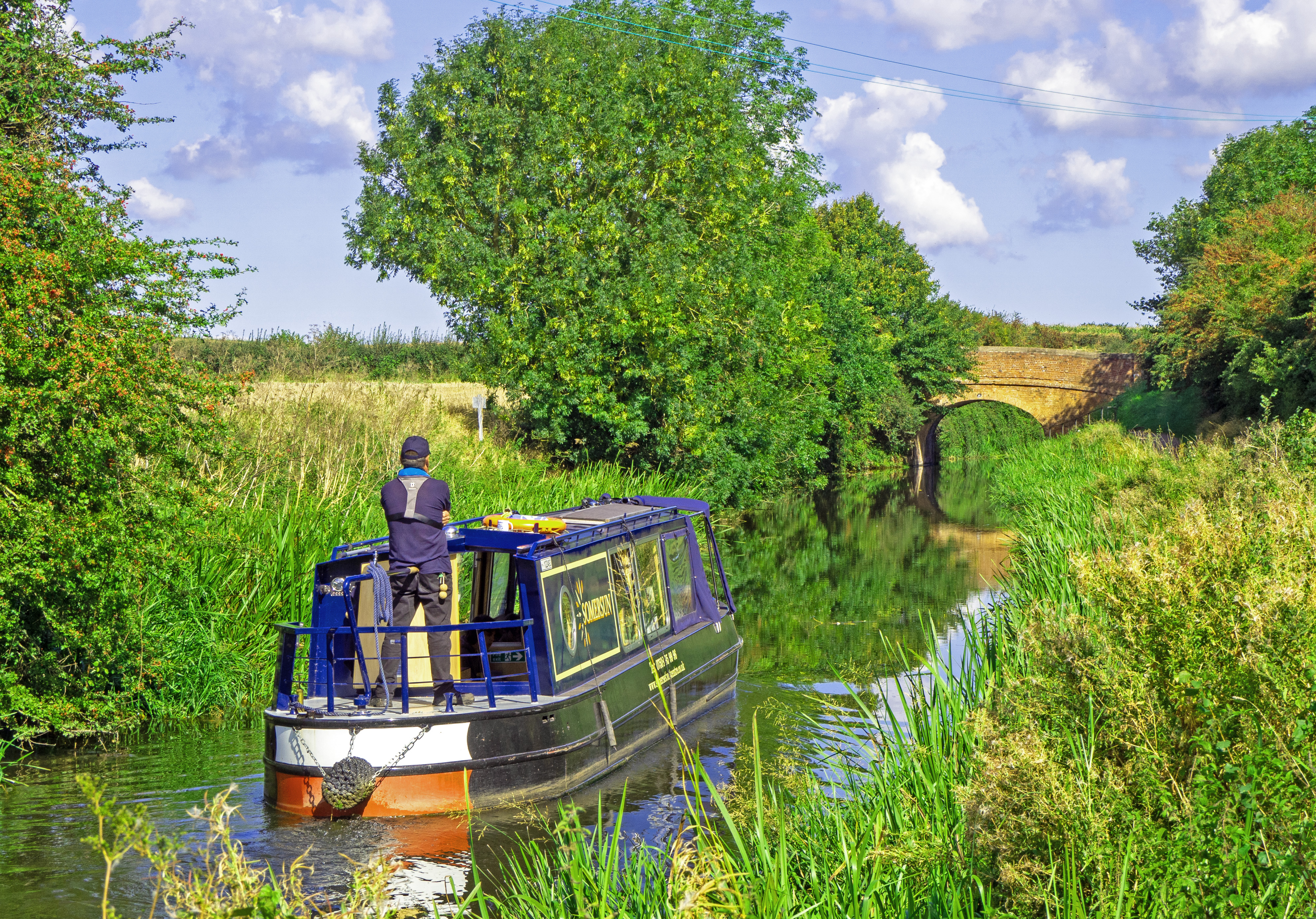 canal boat on canal with man steering and tees and green fields with bridge