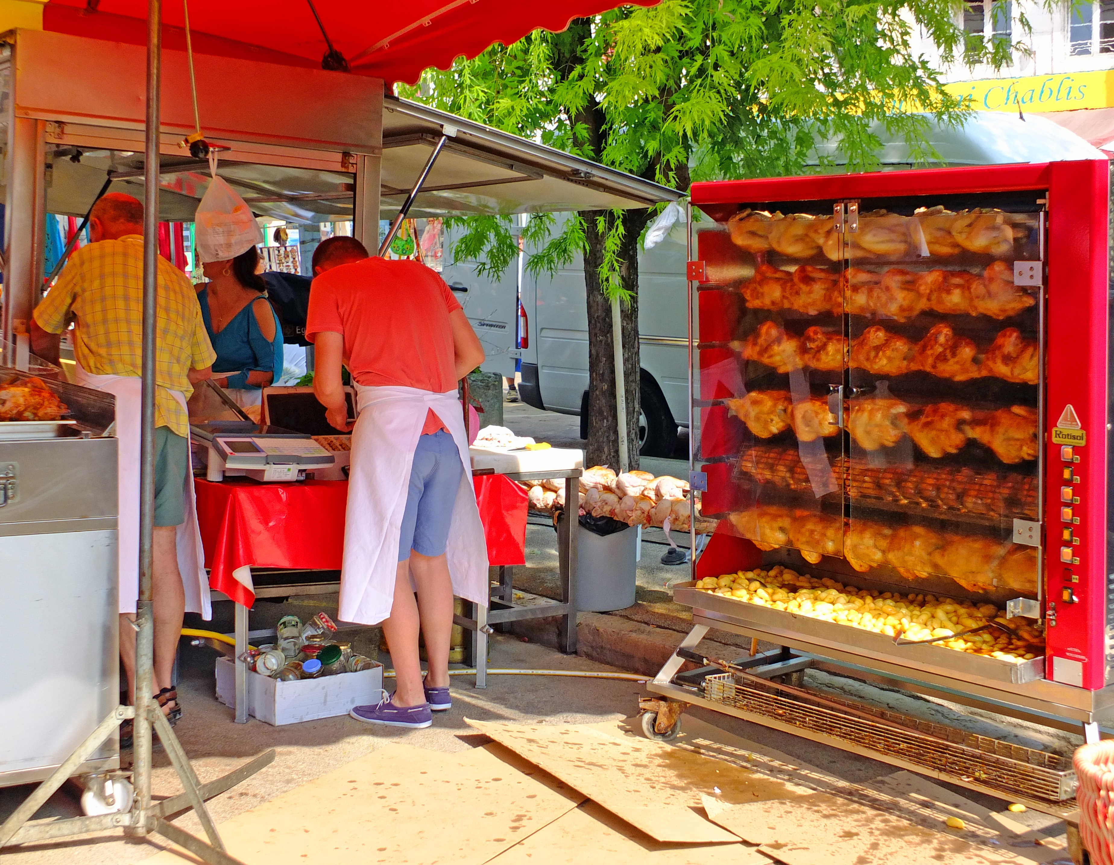 roast chicken and potatoes cooking on spit with people under canopy on market with tree