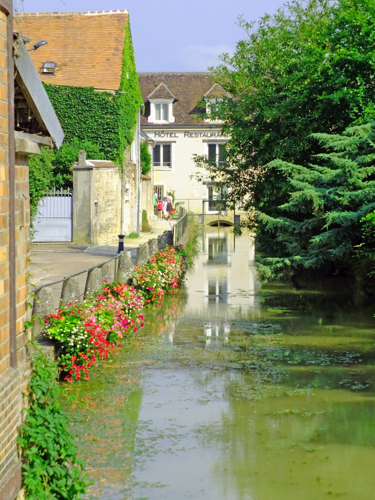 river with flowers and plants on bank with buildings and trees and blue sky