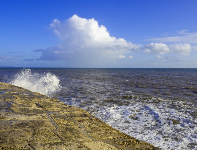 sea waves rise over harbour wall with sea and clouds in blue sky