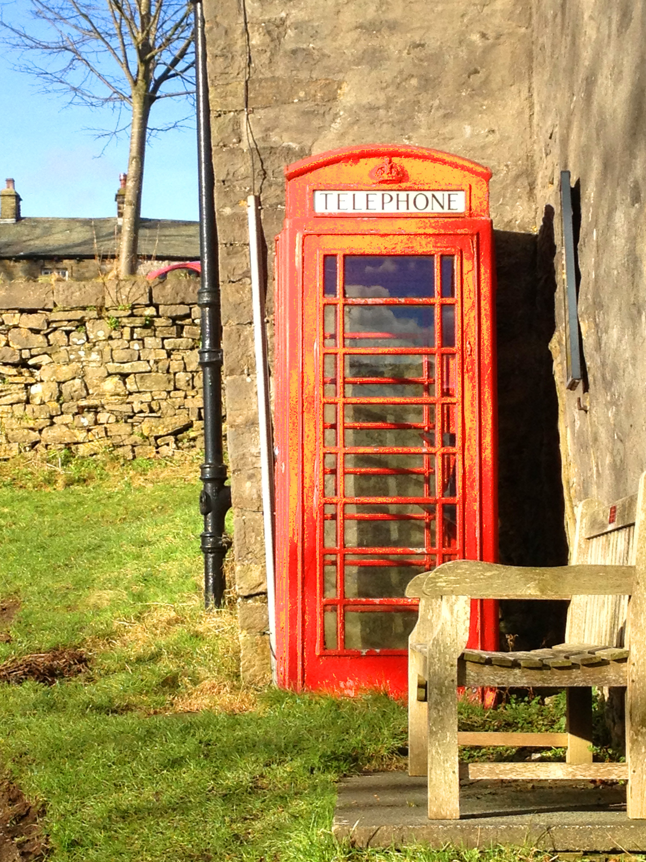 red telephone box with wooden bench and old house with grass and blue sky