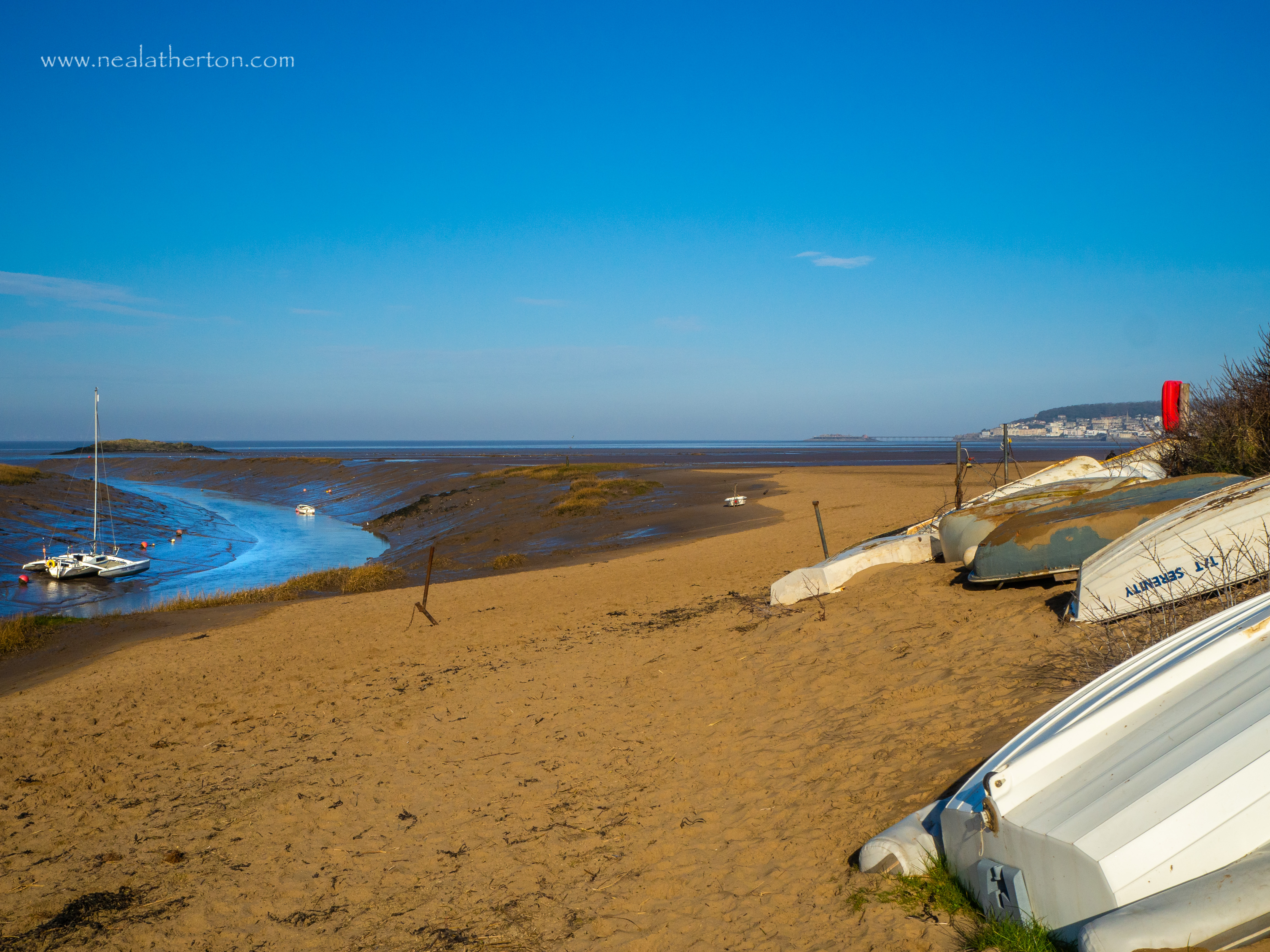 several rowing boats on a sandy bank by a river estuary with the sea and blue sky