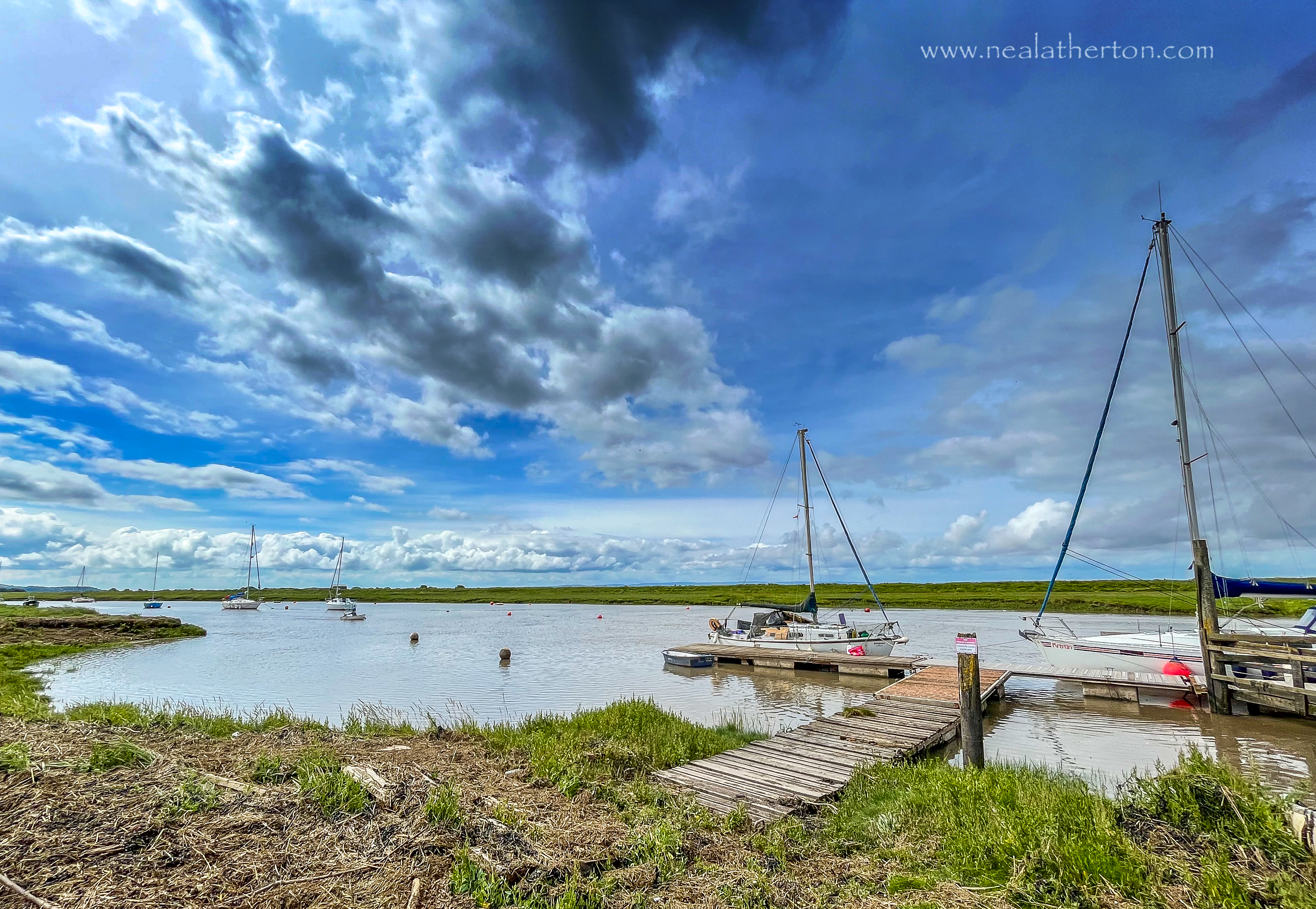 Walkway and jetty with yacht and high river and grassy banks with an unsettled sky
