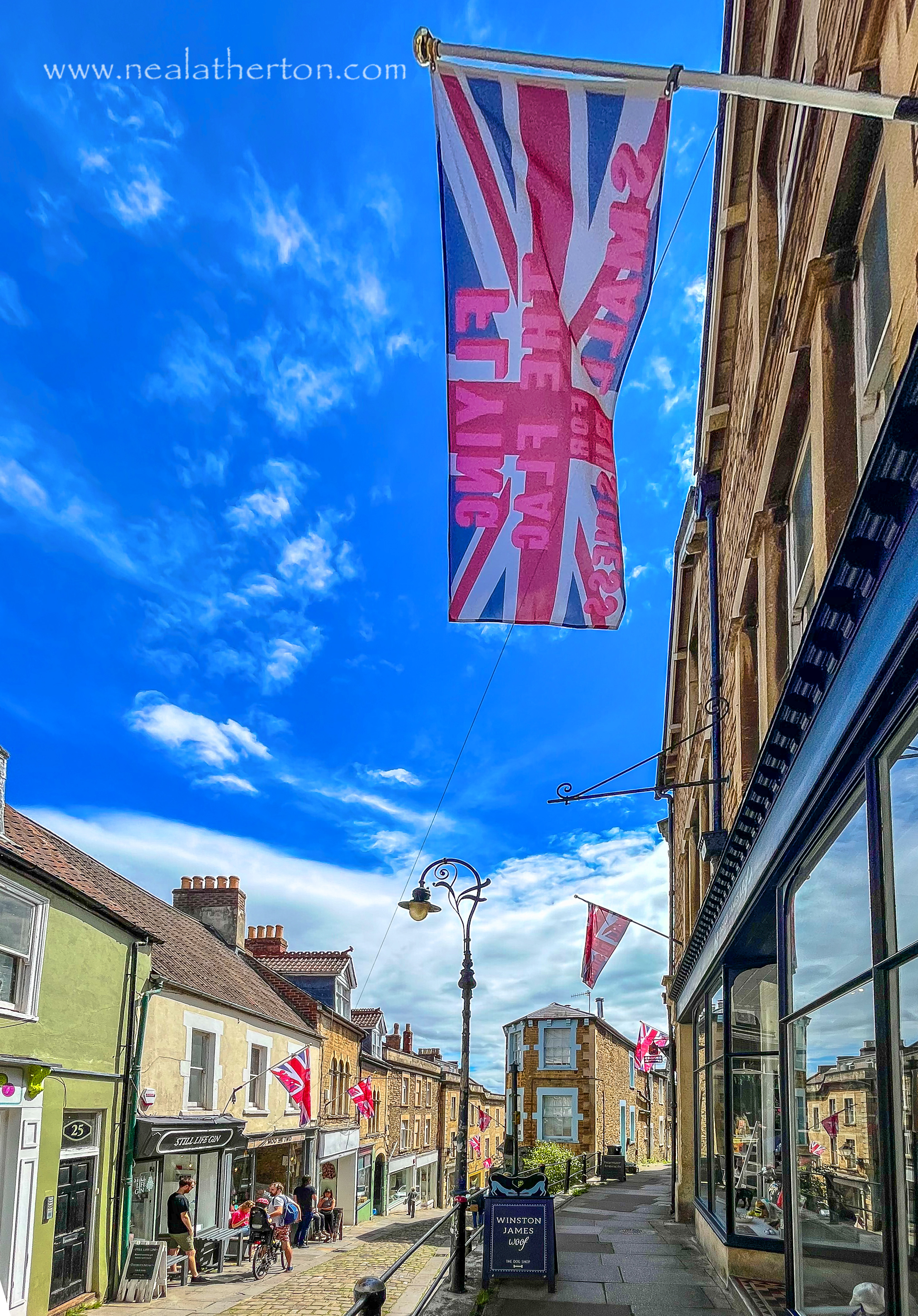 Union jack flag flying from shop on cobbled street with cast irin lamposts and shops under blue sky