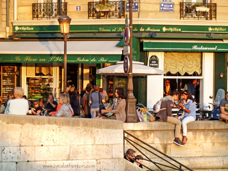 a couple sit close together on river wall with people talking and cafe with green awning and golden stone buildings