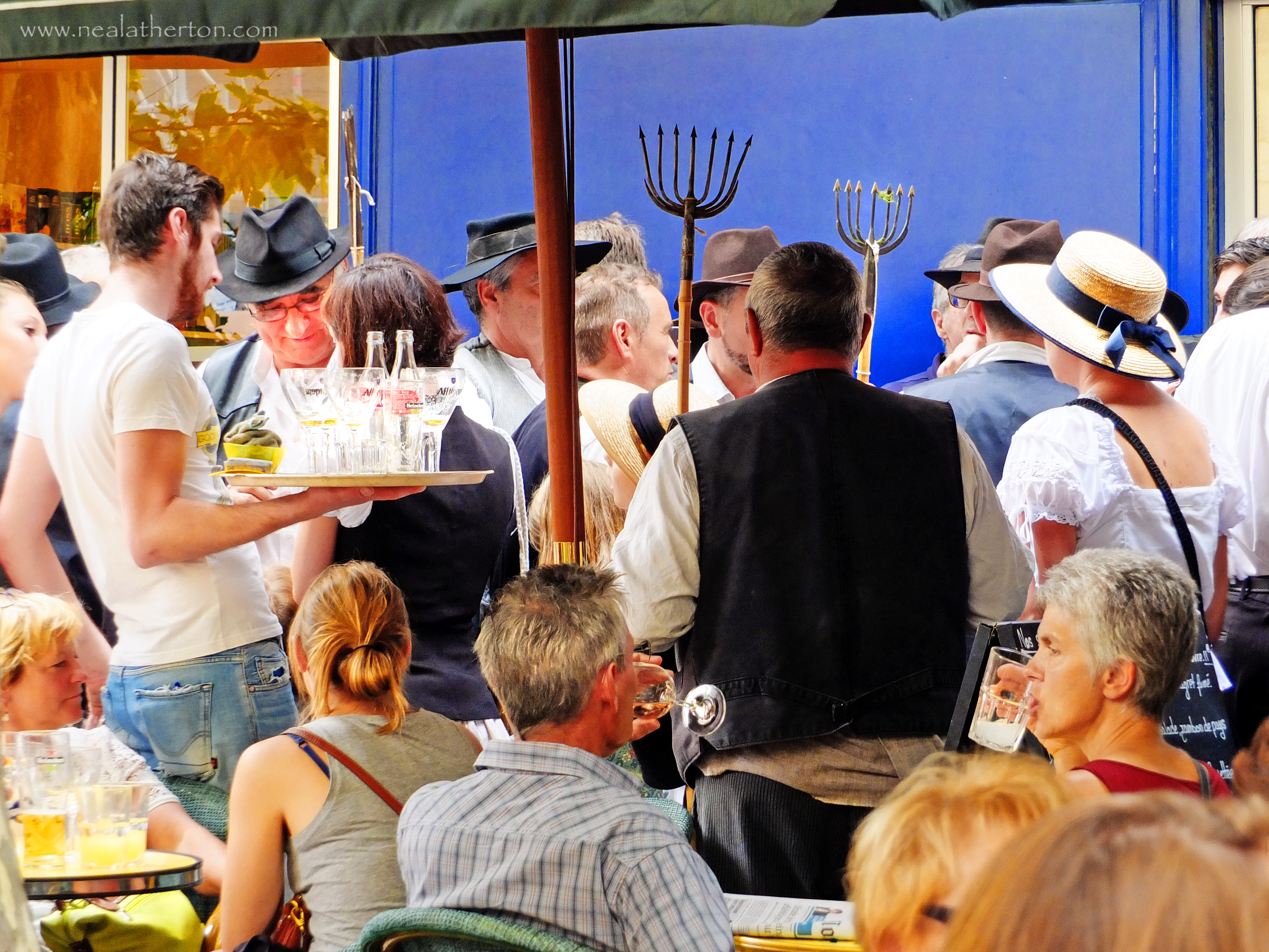 waiter carrying bottles on terrace with people drinking at tables and men carrying farming tools