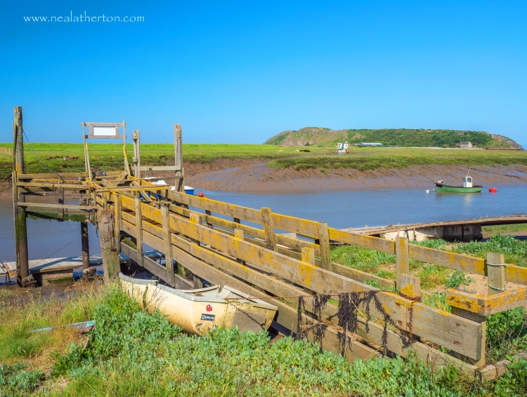 Broken rowing boat by a wooden jetty on an estuary with grassy fields and island and blue sky