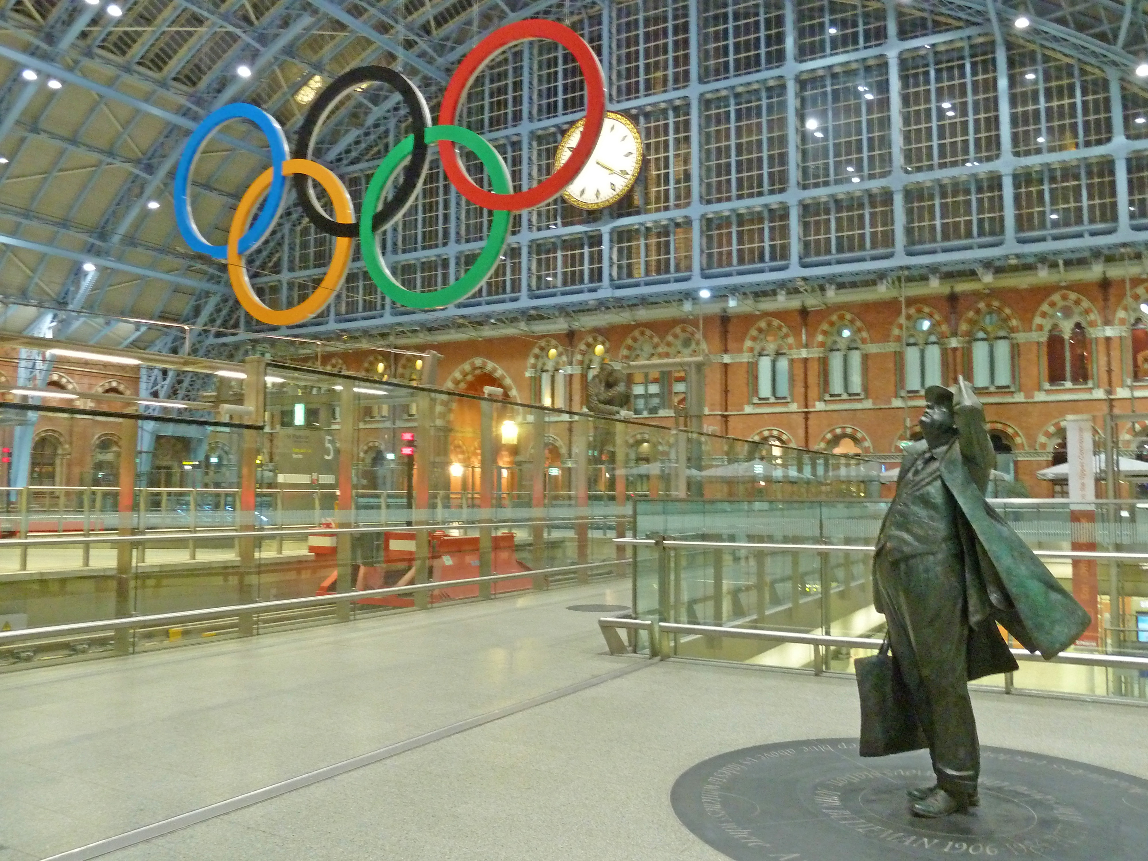 statue of man with bag in front of olympic rings in train station with clock