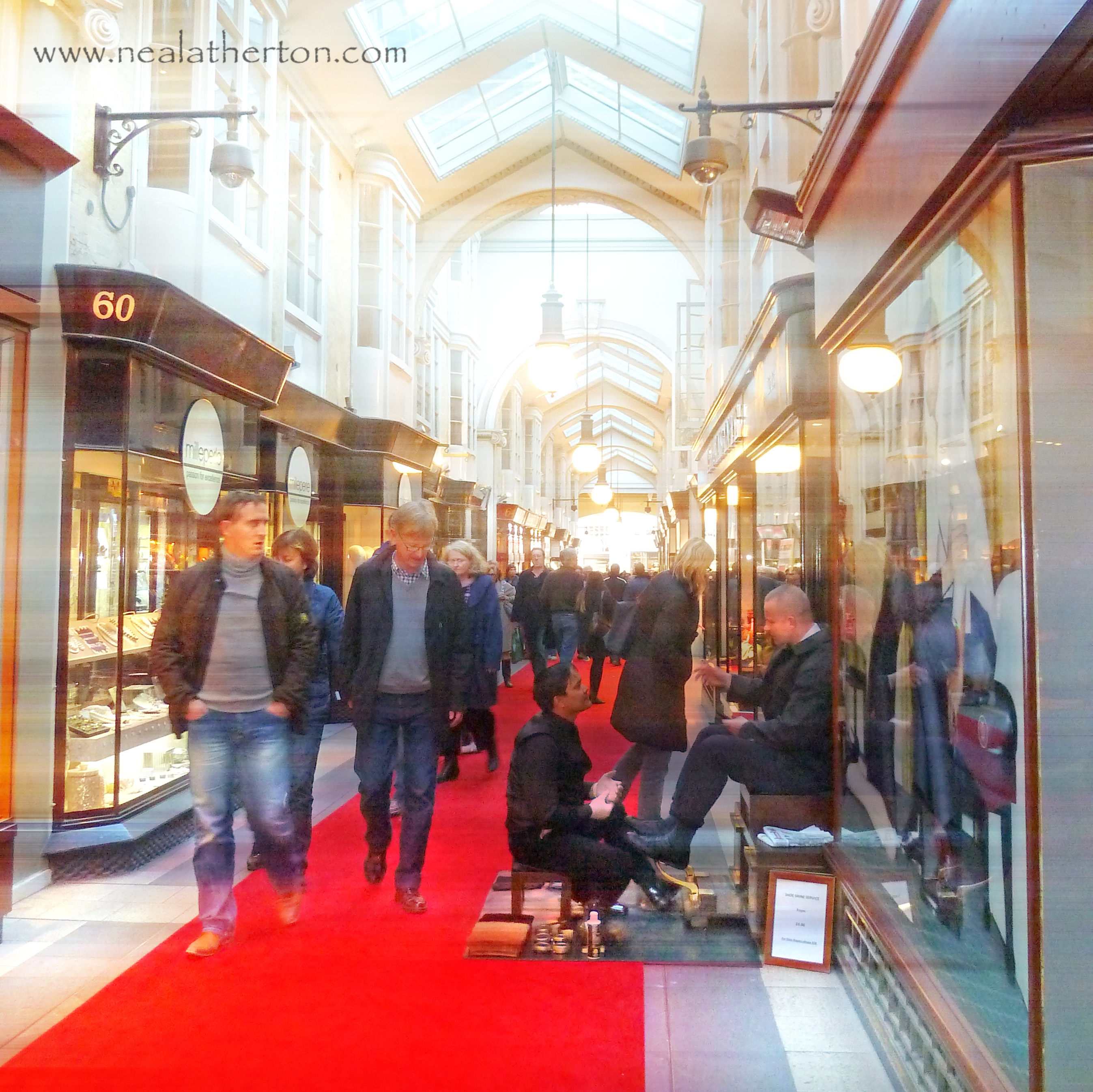 man shines shoes of another man as people walk in arcade of shops with glass ceiling and red carpet