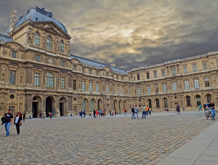people on cobbled courtyard in front of period architecture with threatening sky