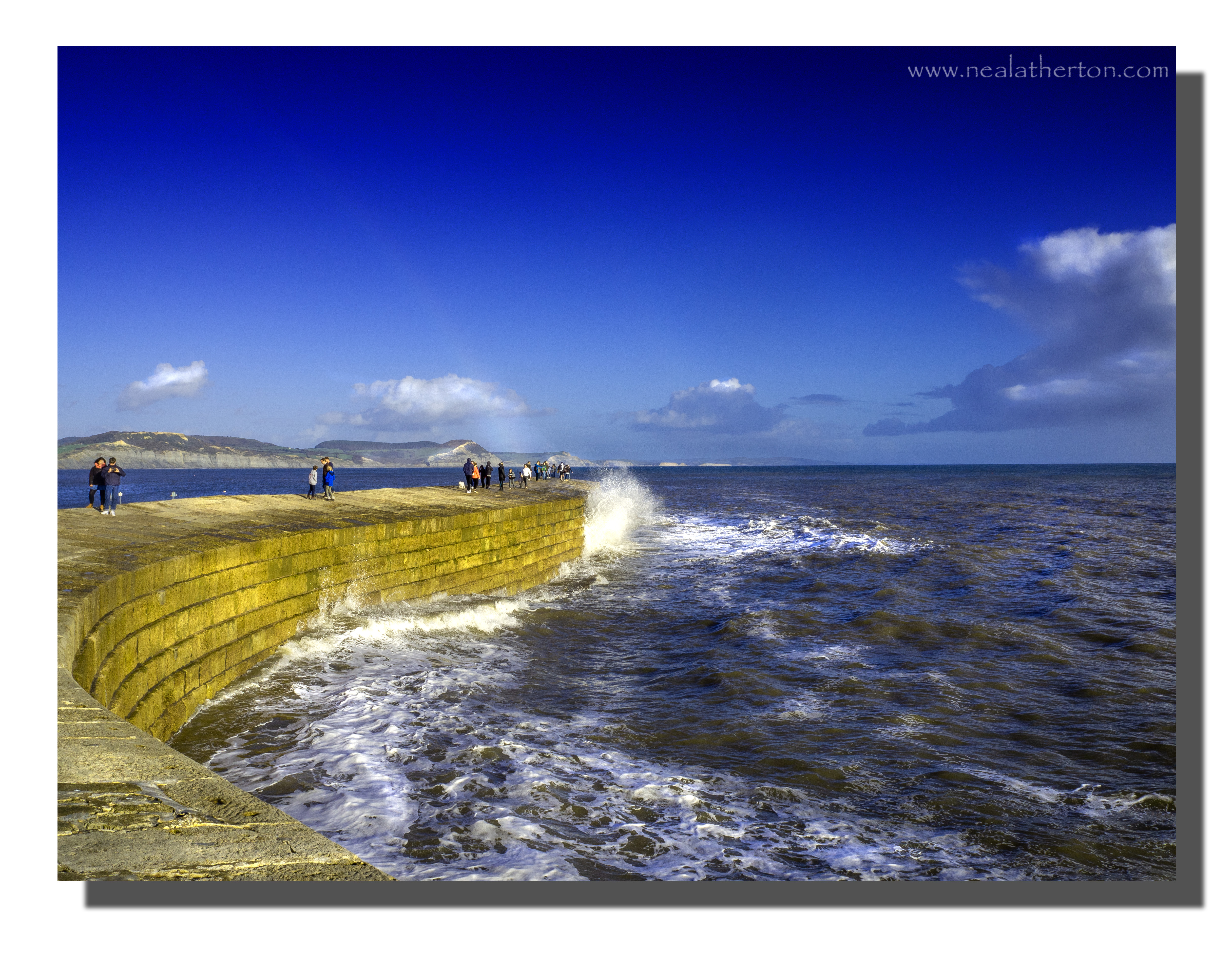 Heavy sea waves hitting harbour wall with people walking and blue sky