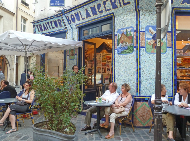 people sit at tables to drink on terrace of shop on cobbled street
