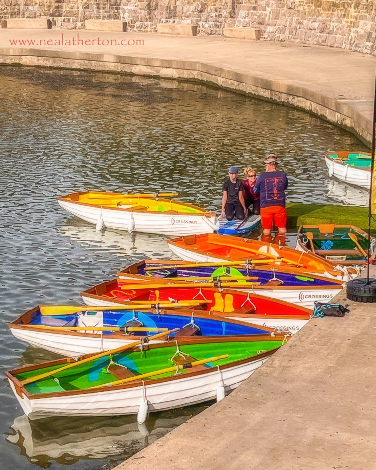 eight different coloured boats on a lake with three people and a stone wall and walkway