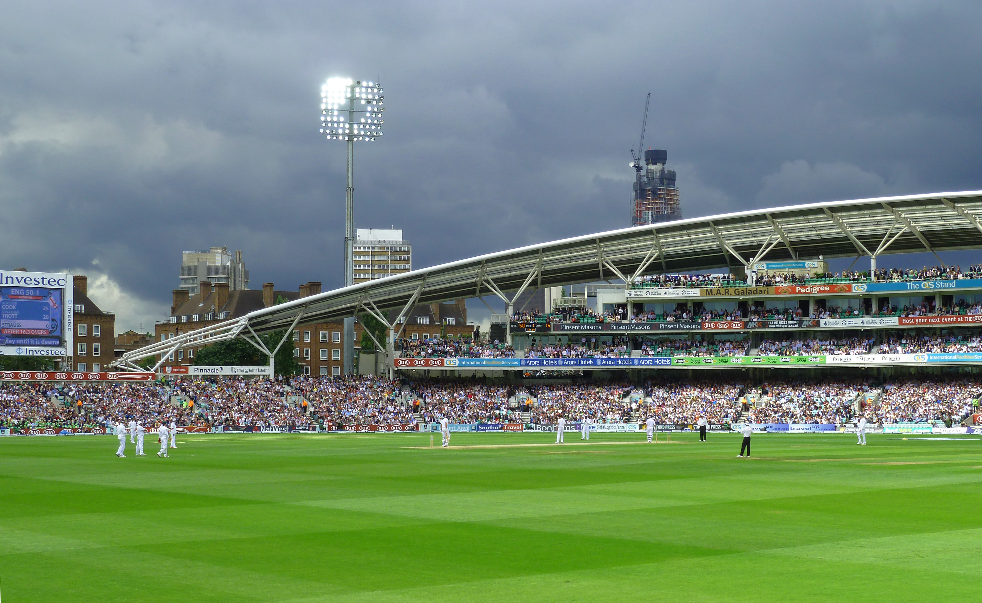 men playing cricket on large ground with people in large stand with floodlights and grey sky