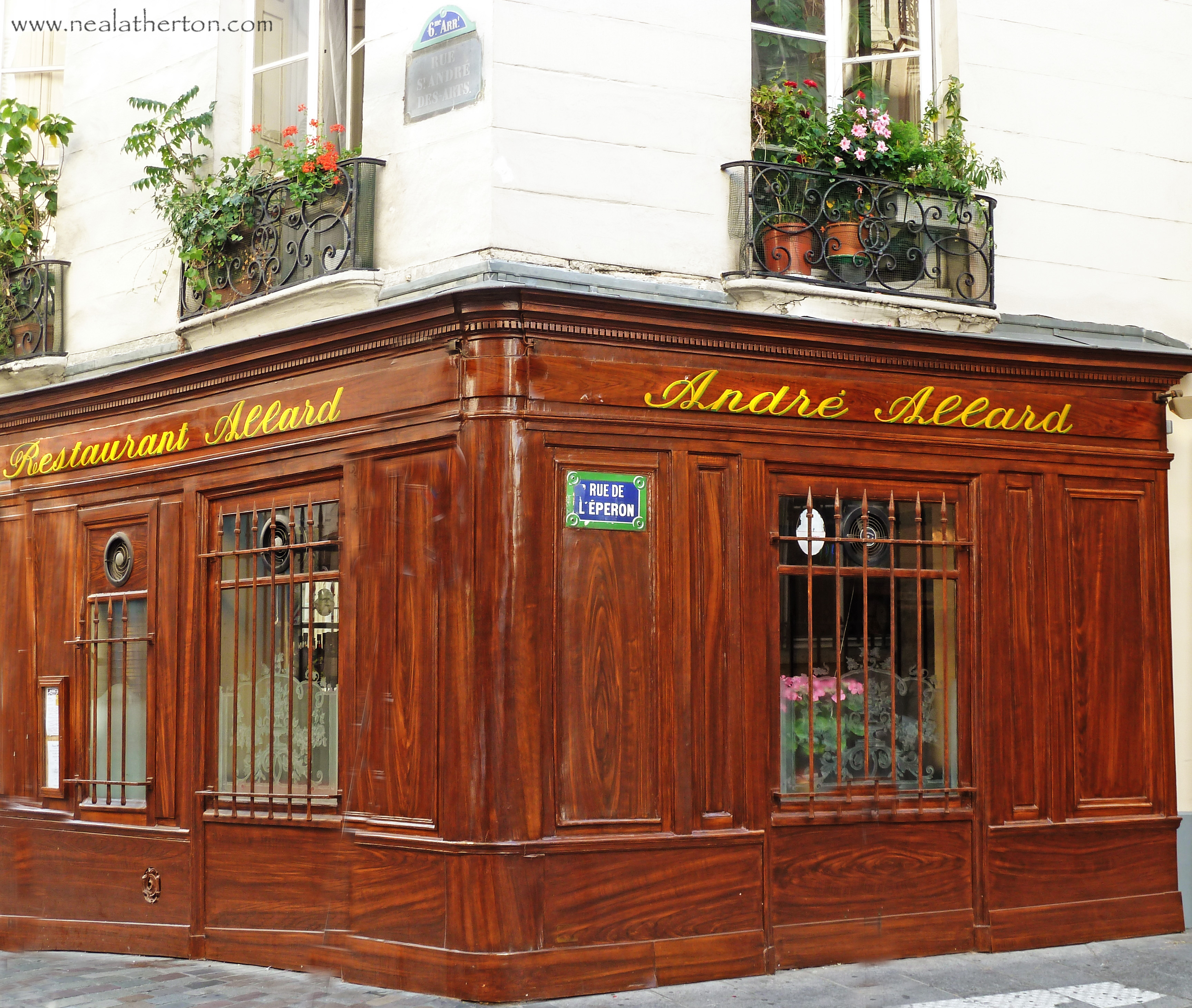 wooden front on restaurant with gold lettering and street sign with plants on balcony