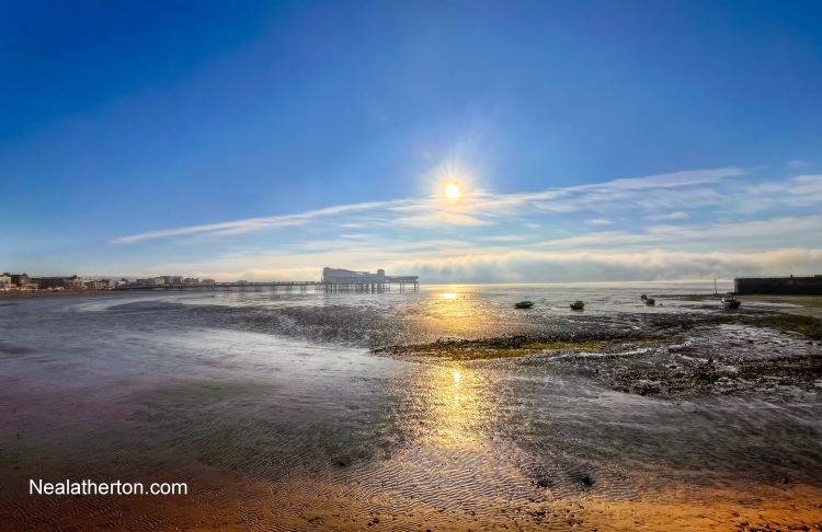 sun reflecting on water on beach with pier and sea mist with baots in foreground