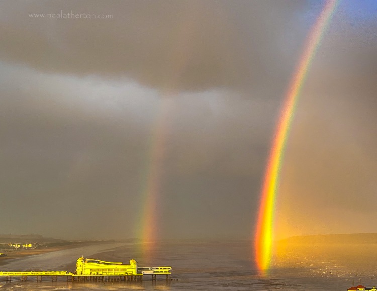 two rainbows brightly coloured with pier and sea in front of stormy black clouds