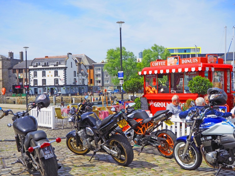 four motorcycles on waterfront by mobile cafe with people eating and buildings