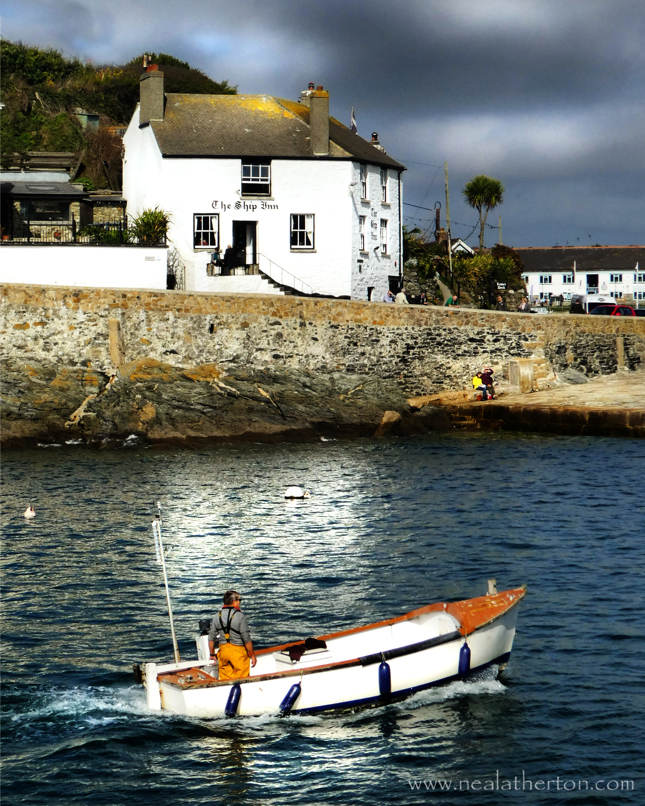fishing boat goes into harbour with pub and harbour wall with stormy sky