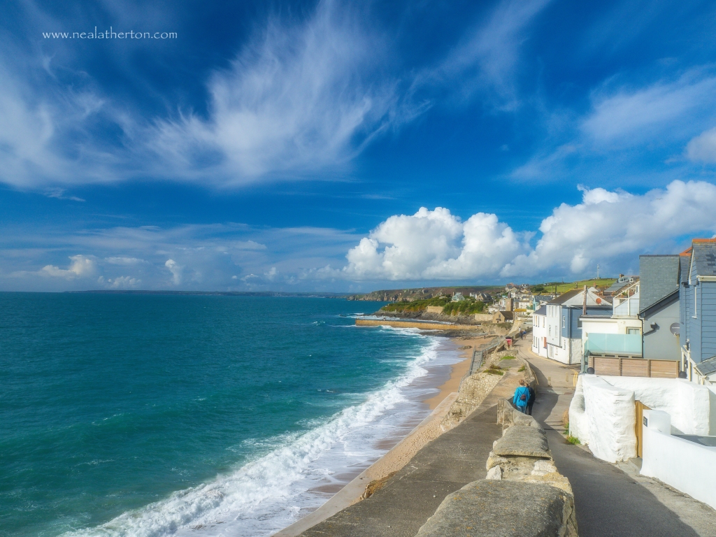 path leads past white painted cottages on cliff overlooking beach and sea with town beyond