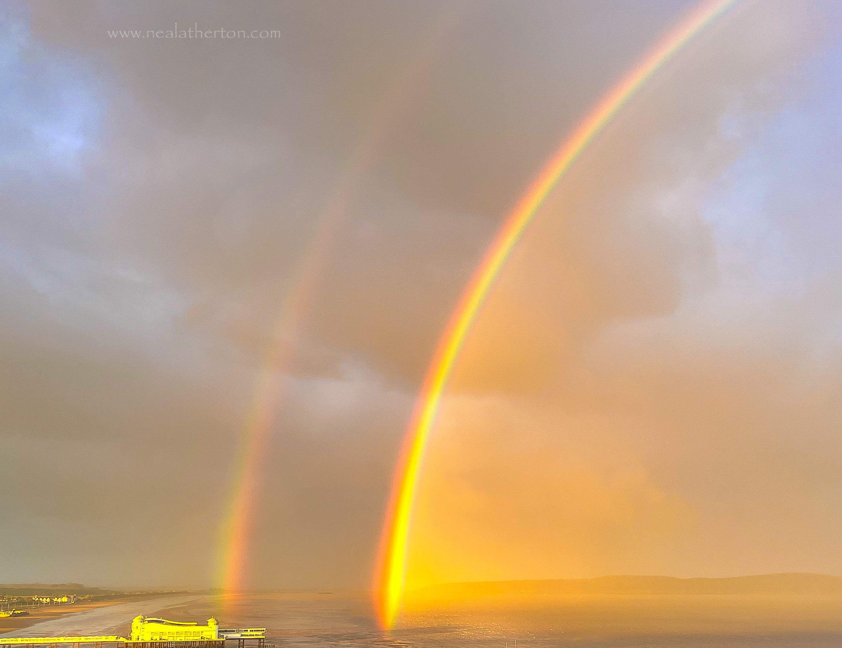 Pier on bay with two rainbows brighly colured against a stormy sky