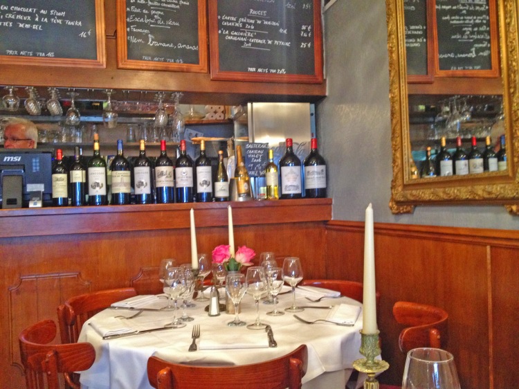 Table in restaurant with candles and glasses with wine bottles on bar and chalkboard menu
