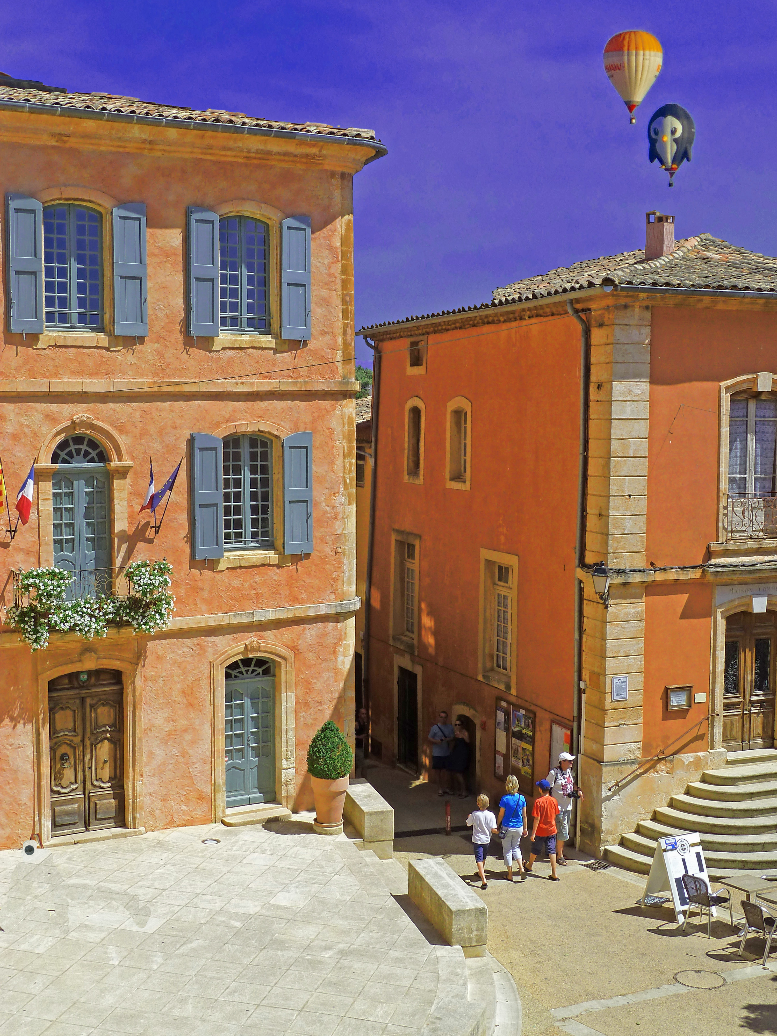 Two hot air balloons above old terracotta coloured buildings in village square with people walking