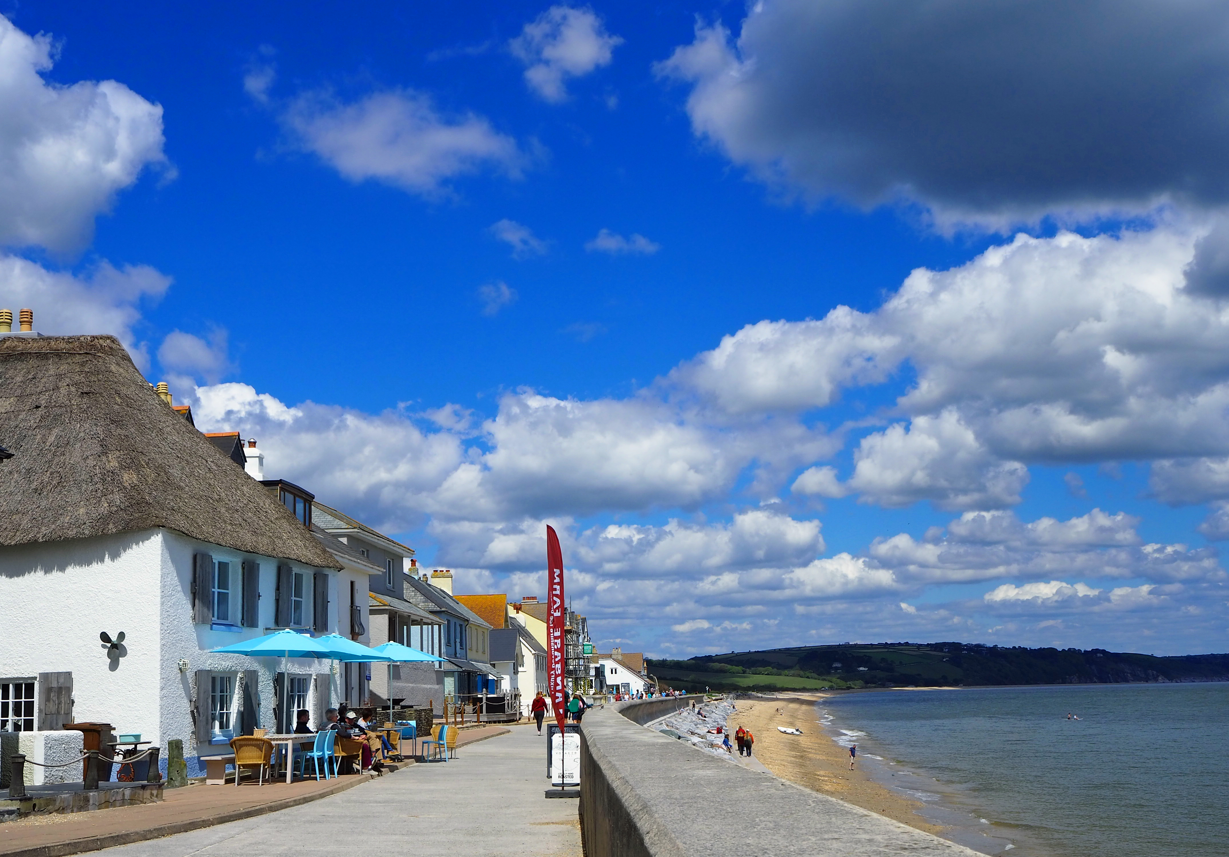 thatched houses on sea front with sea wall and tables and chairs with parasol along beach