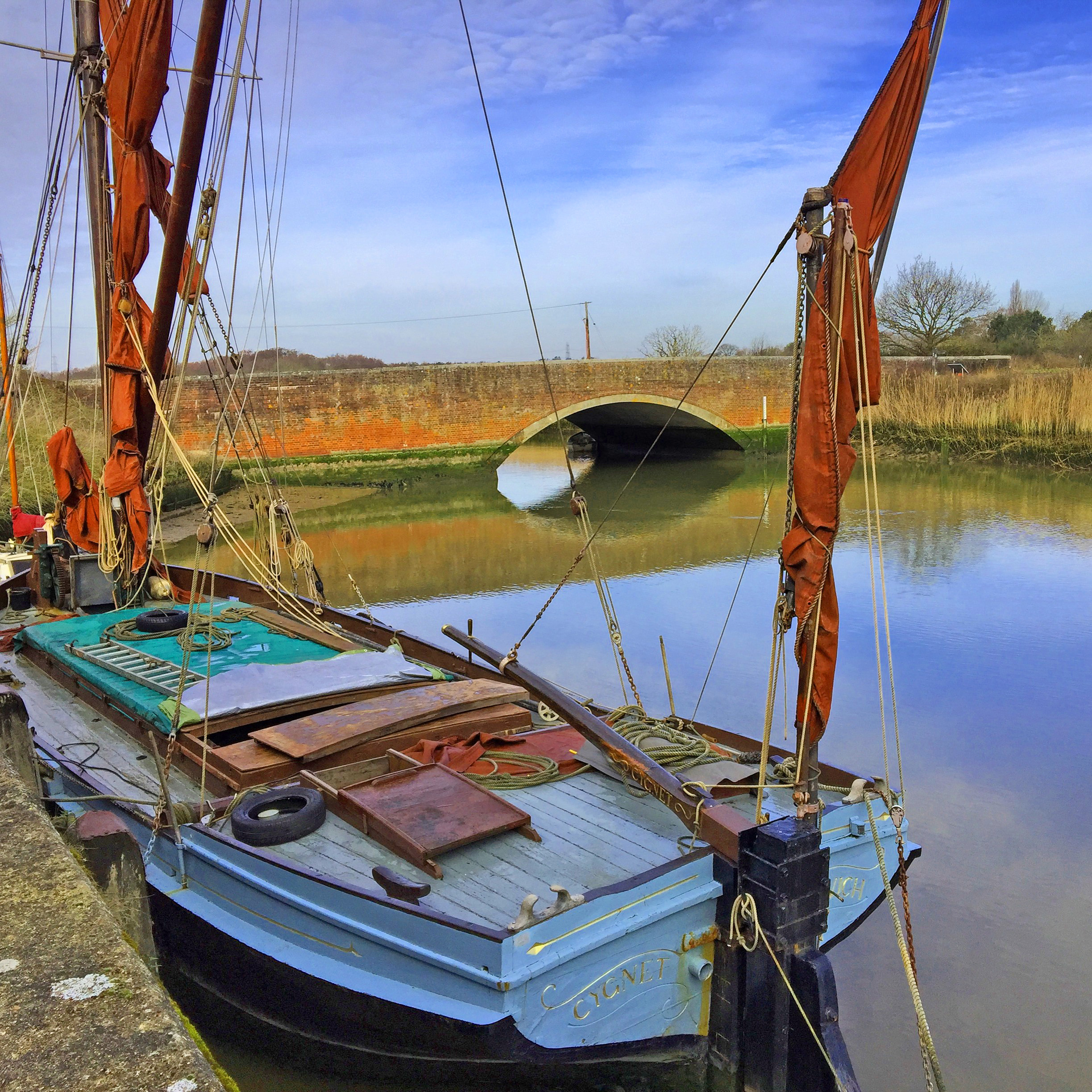 boat with red sails on river with brick bridge and blue sky