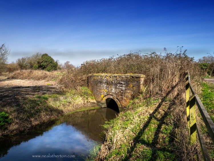 bridge over stream with grassy bank and field with wooden fence and path with trees