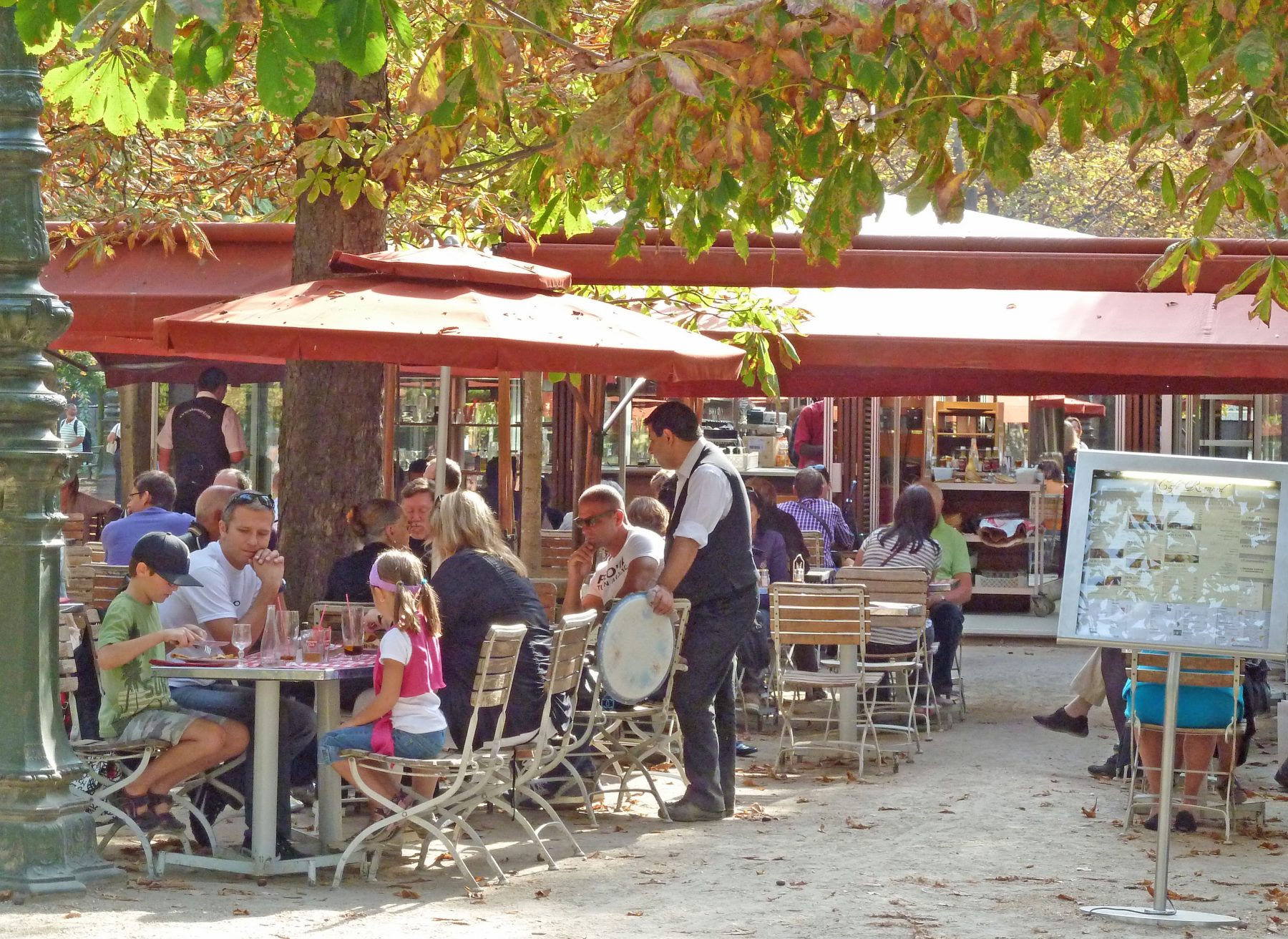 waiter talks to customers at tables in cafe in garden under trees