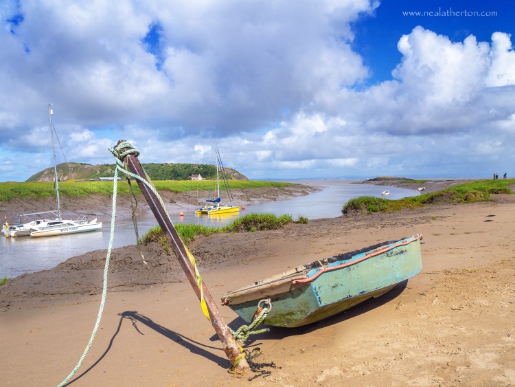 Old little boat by metal post in sand with river and yachys and hill in front of cloudy summer sky