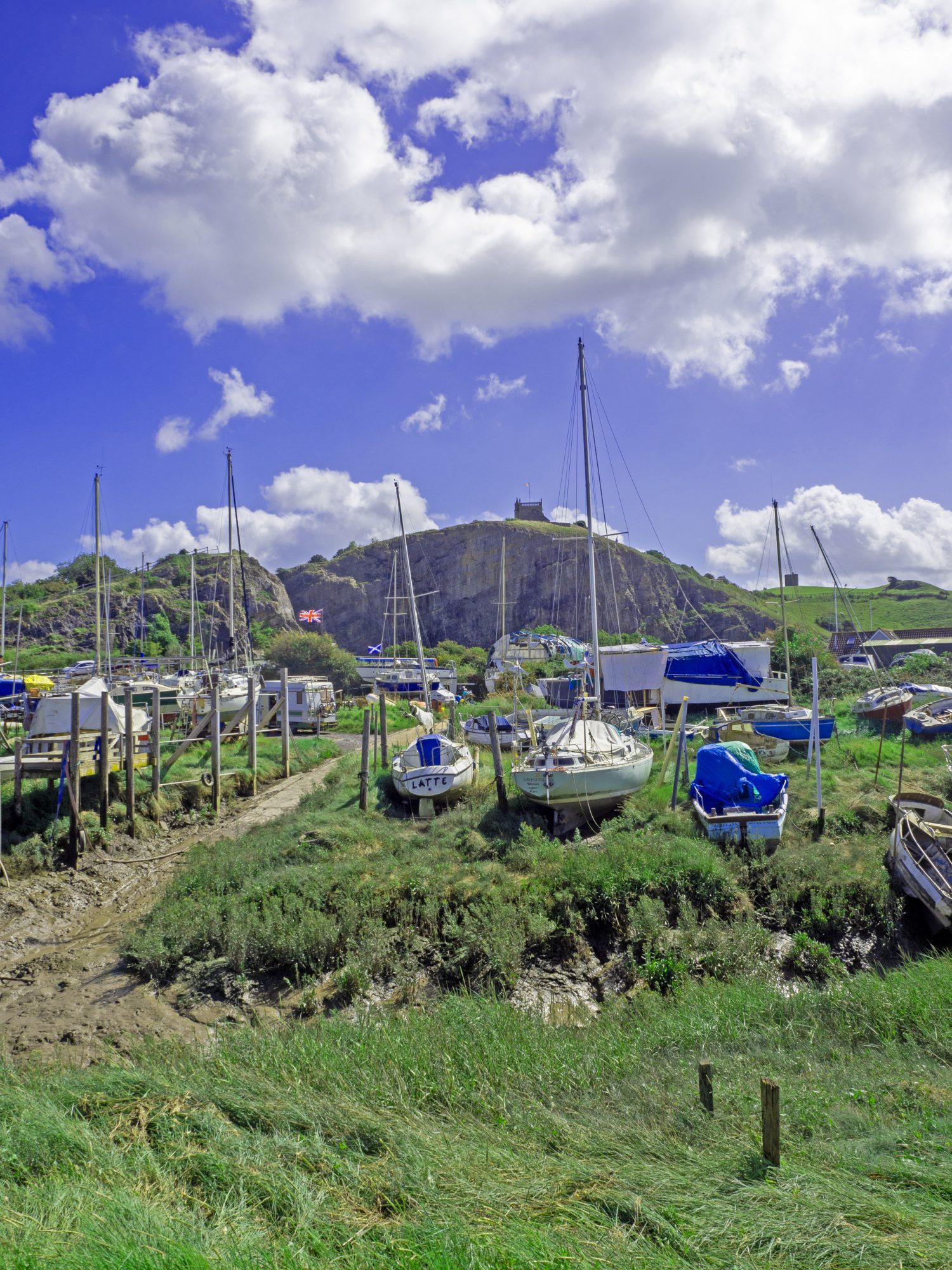 yachts and boats on bank at boatyard withgrassy bank and quarry with church on top