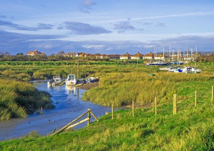 yachts and boats on river at boatyard with grassy bank and fence and blue cloudy sky over houses