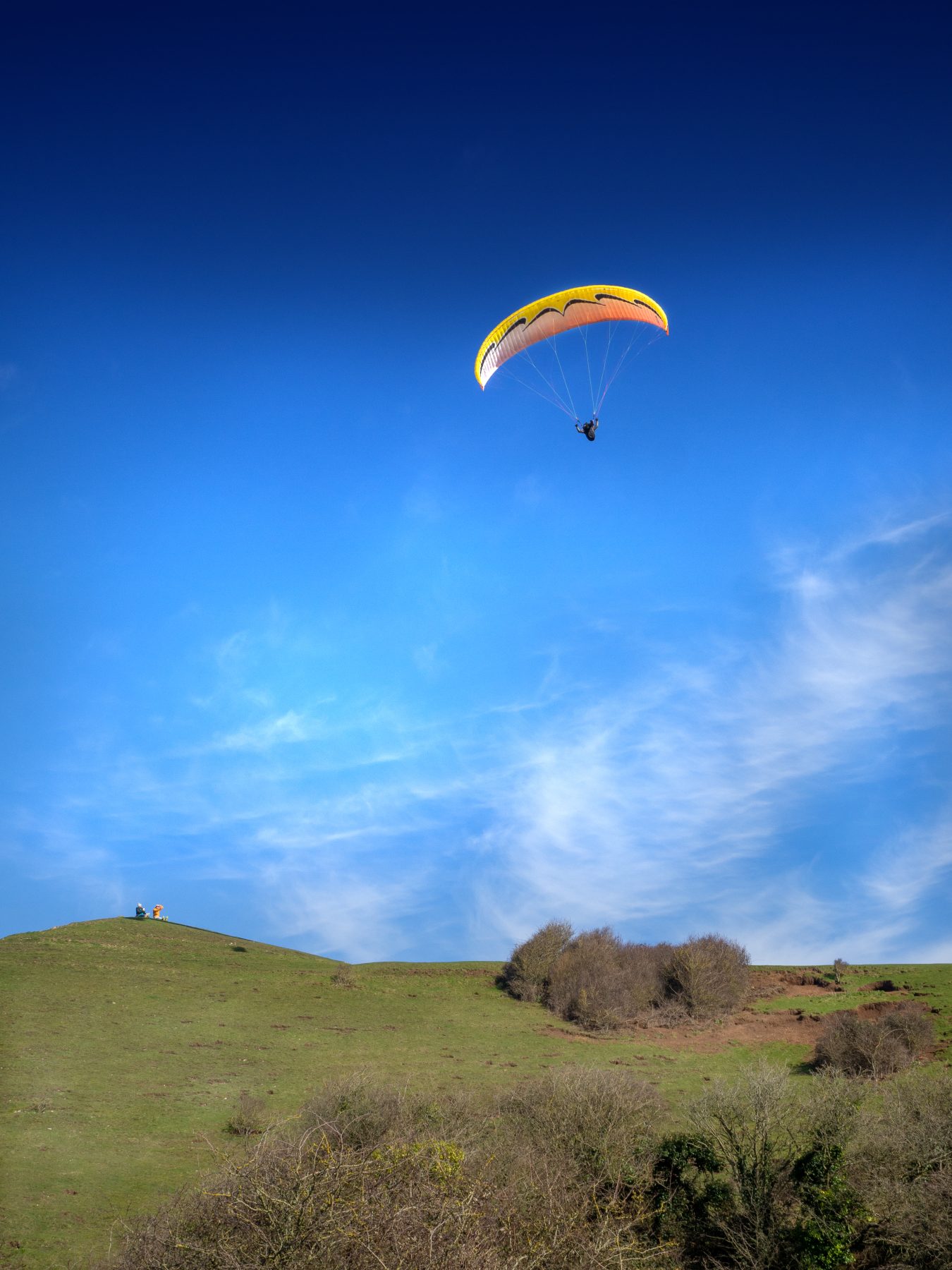 yellow handglider over grassy hill and bushes with blue sky