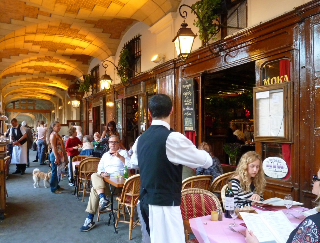 waiter serves at table on terrace by old wooden fronted cafe under stone archway