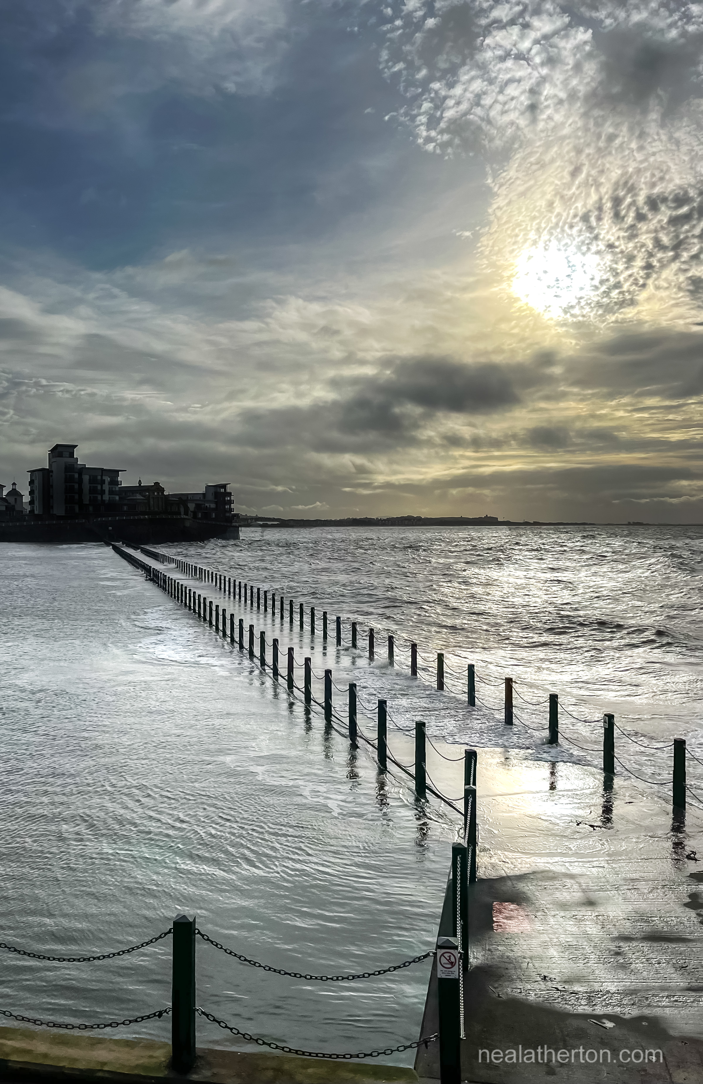 Sun behind winter clouds in dark sky with walkway over the sea to an island