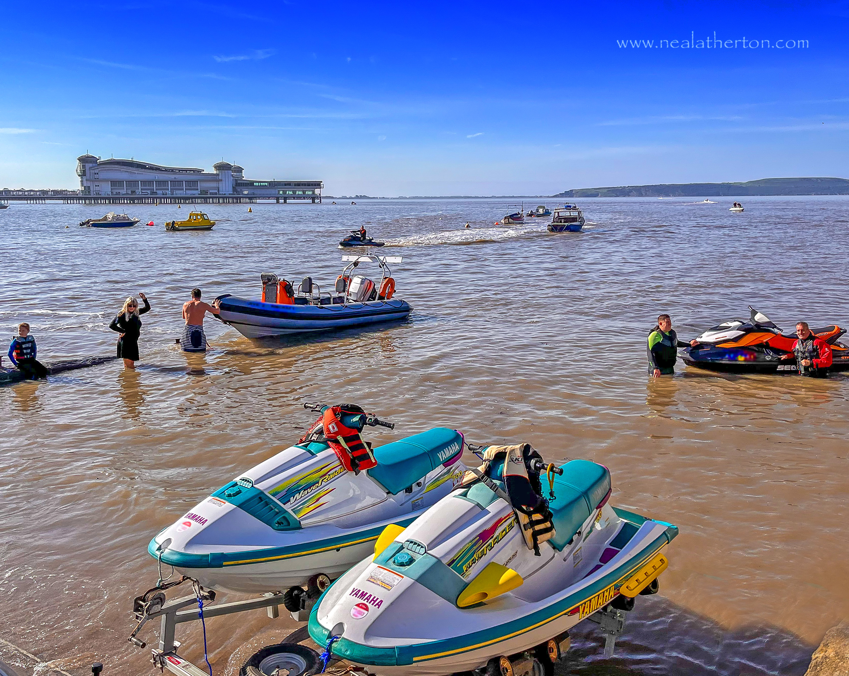 Four jet ski and other boats with people in the water with Weston pier behild under summer blue sky