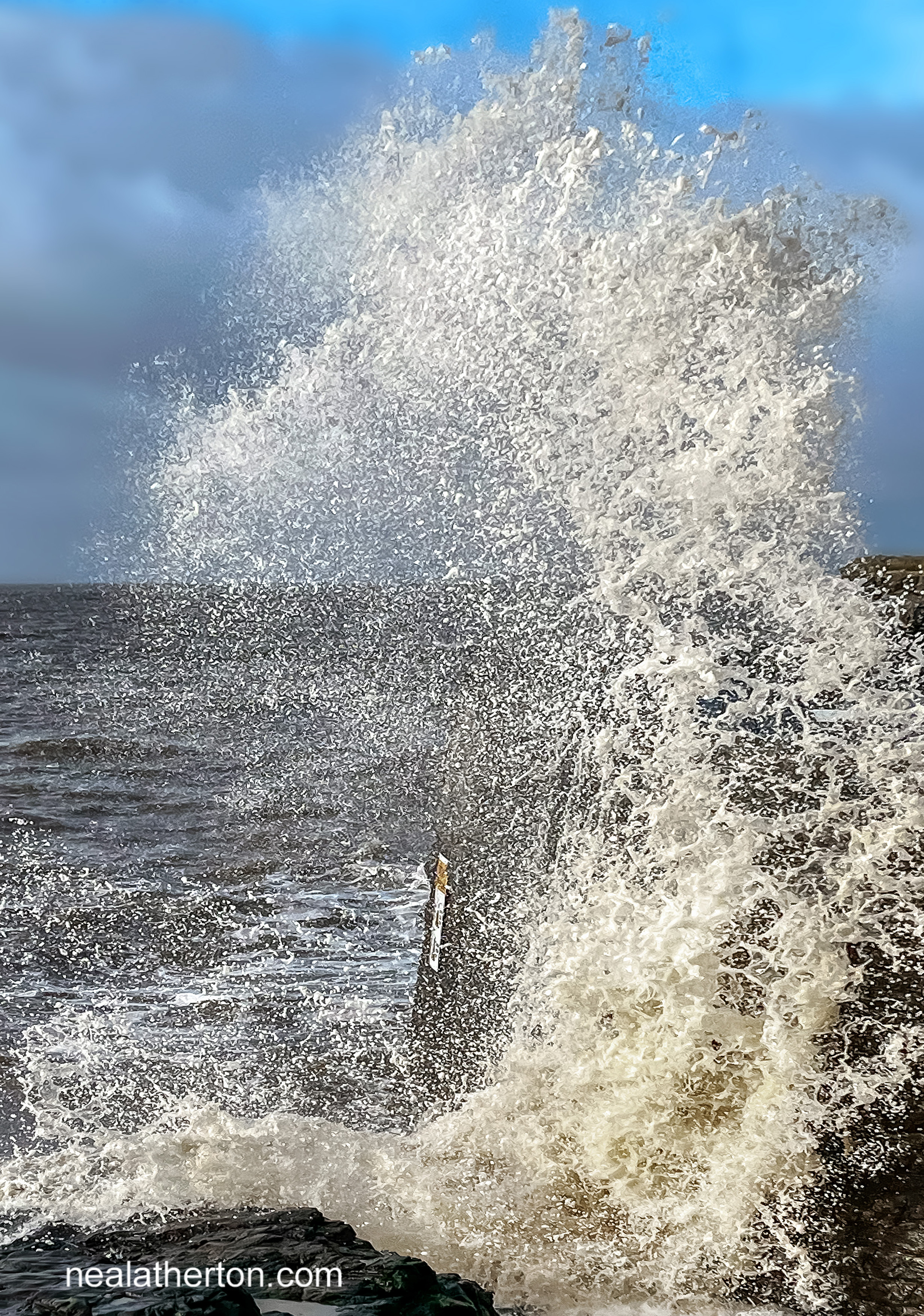 sea splashes up against the sea wall in Weston with blue and cloudy sky behind
